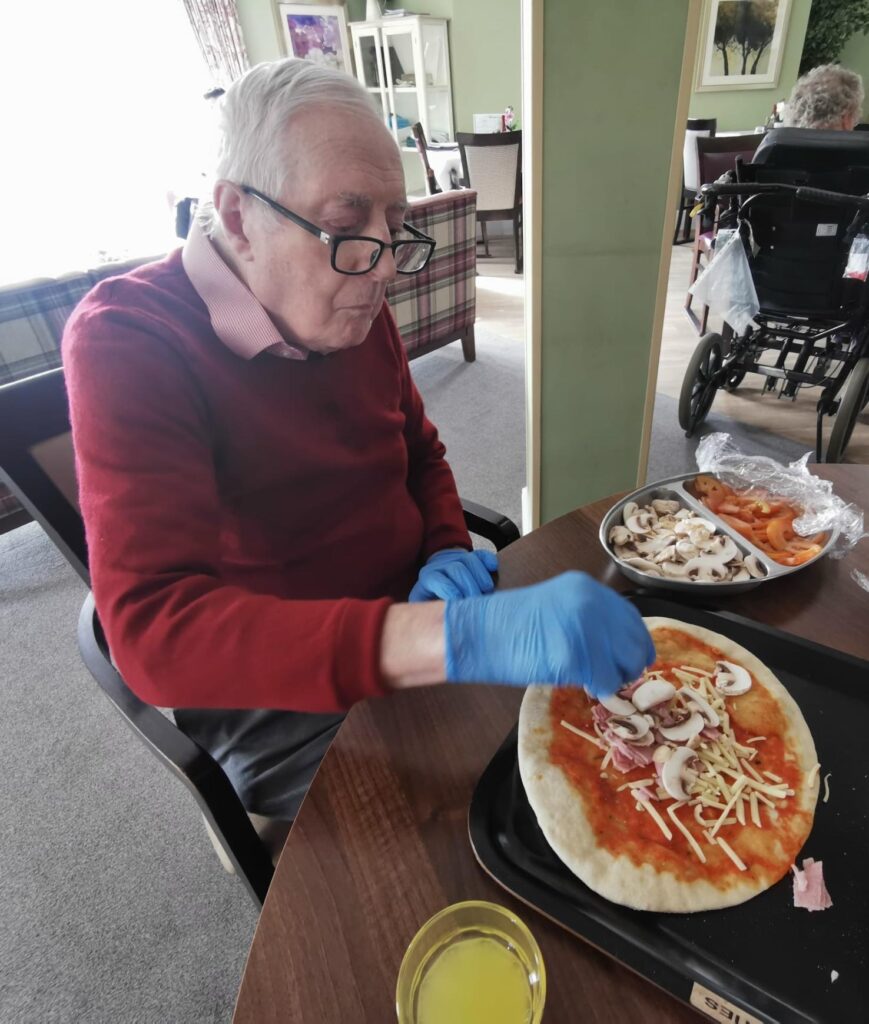 resident with blue gloves on decorating a pizza with mushroom and ham