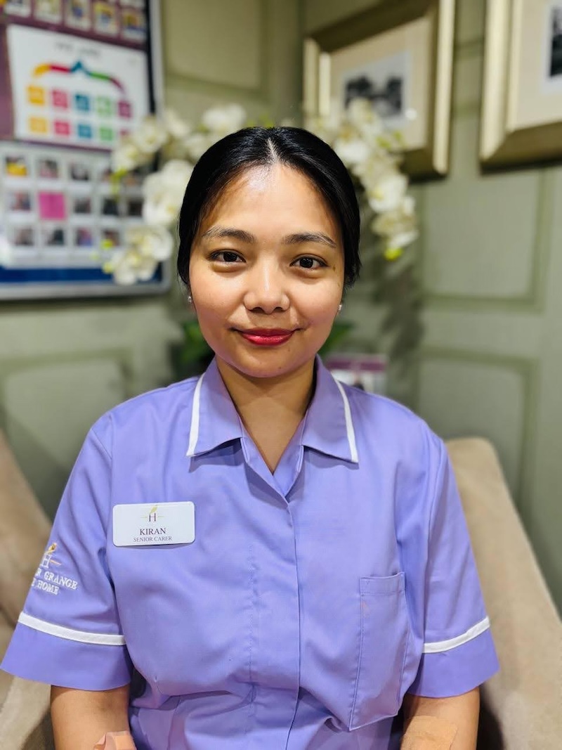 Woman wearing a light purple senior carer uniform sat in an armchair smiling for a headshot