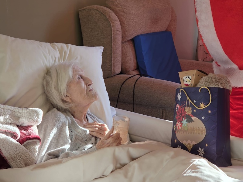 elderly lady in bed holding a christmas present smiling at santa