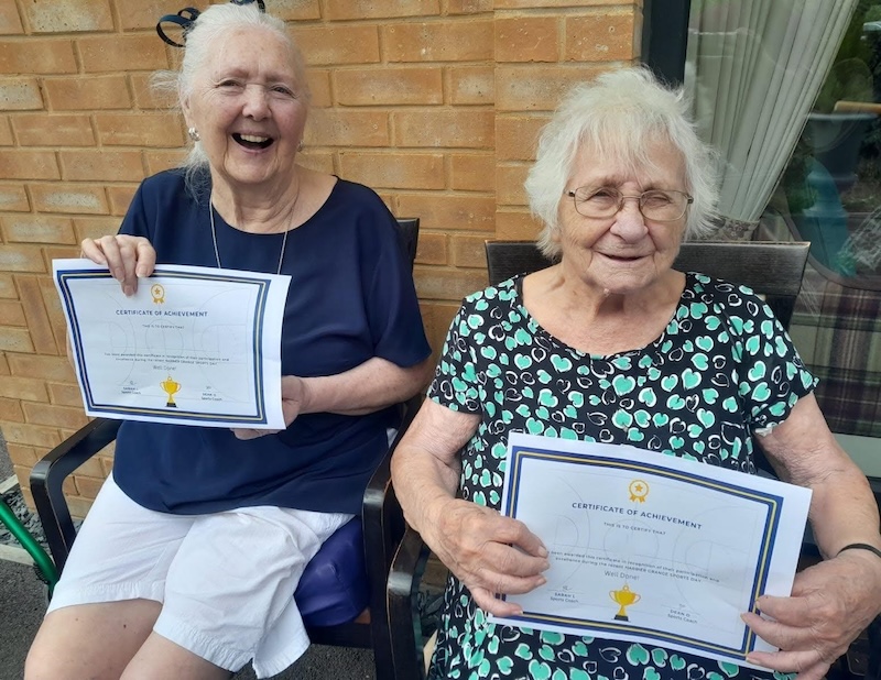 2 elderly females sat in the garden smiling holding certificates